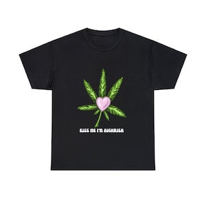 Könnte beinhalten: Schwarzes T-Shirt mit einem grünen Marihuana-Blatt-Design und einem pinkfarbenen Herzen in der Mitte. Der Text "Kiss Me I'm Highrish" ist unter dem Blatt aufgedruckt.