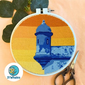 May include: A cross-stitch embroidery hoop featuring a blue and yellow design of a building with a domed roof, set against a gradient yellow and orange sky. The hoop is white, and a pair of decorative scissors rests nearby.
