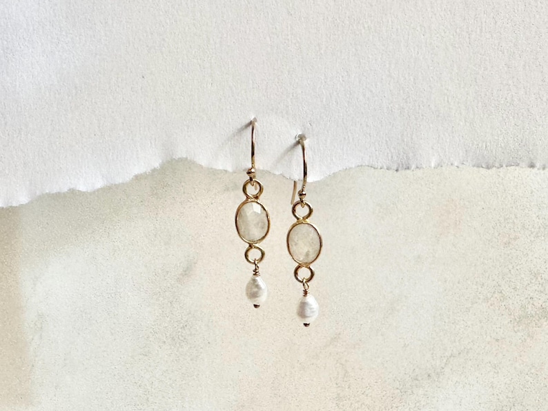 May include: A pair of gold earrings with white oval-shaped stones and a single white pearl dangling from each earring.