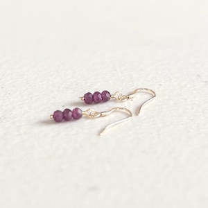 May include: A pair of dangle earrings featuring three small, faceted, deep purple beads strung on thin gold-toned wire. Each earring is attached to a simple gold-toned hook ear wire, creating a delicate and minimalist design for the ears.