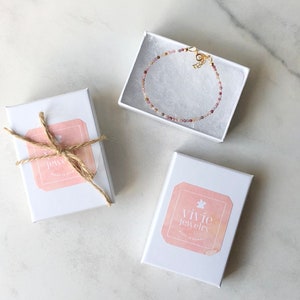 May include: Three white gift boxes with pink and white labels. The boxes are on a white marble surface. One box has a brown twine bow and a label that says "Vivie Jewelry". Another box has a label that says "Vivie Jewelry". The third box contains a delicate pink and gold beaded bracelet.