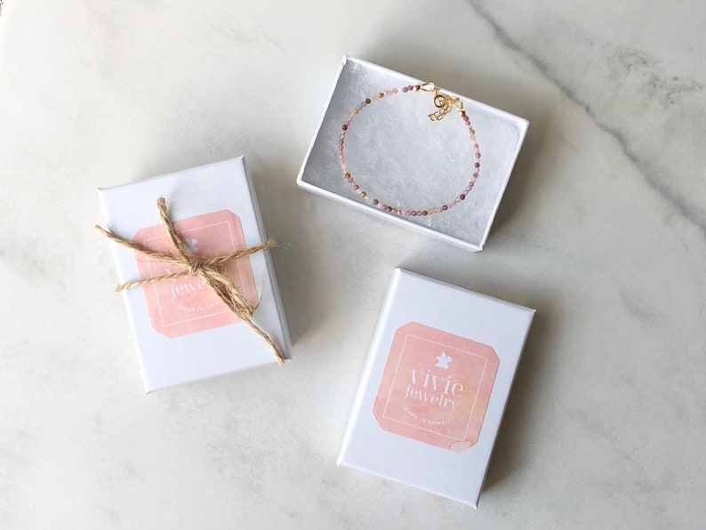 May include: Three white gift boxes with pink and white labels. The boxes are on a white marble surface. One box has a brown twine bow and a label that says "Vivie Jewelry". Another box has a label that says "Vivie Jewelry". The third box contains a delicate pink and gold beaded bracelet.