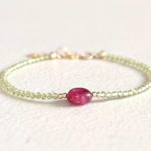 May include: A delicate bracelet featuring a single oval-shaped magenta gemstone centered between small, faceted light green beads. The design includes tiny gold-toned metallic spacers and a gold-toned chain closure at the back of the piece.