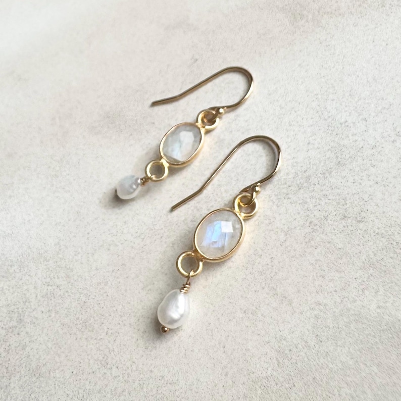 May include: A pair of gold earrings with a teardrop pearl and a moonstone oval. The earrings have a simple design and are perfect for everyday wear.
