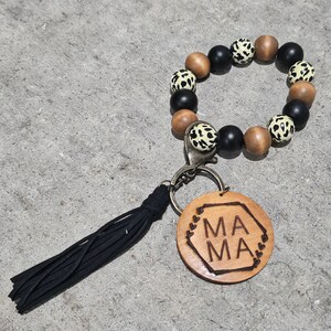 May include: A wooden keychain with a black tassel and a round wooden charm that says "MAMA" in a hexagon shape. The keychain is made of wooden beads with a black and white leopard print design.