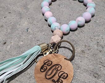 May include: A light blue and pink silicone beaded bracelet with a wooden charm and a tassel. The charm has a monogram engraved on it.
