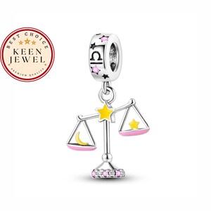 May include: Silver charm with a pink and yellow enamel Libra zodiac symbol. The charm features a scale with a crescent moon and a star on each side. The charm is decorated with small pink and white crystals.