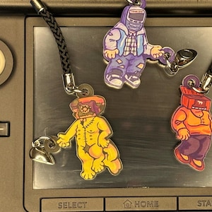 以下が含まれることがあります： 黒い紐の付いたカラフルなキャラクターチャームが3つ。チャームはプラスチック製で、頭が電話の黄色いキャラクター、青と白のシャツを着た紫のキャラクター、頭が赤い箱のオレンジ色のキャラクターが特徴です。