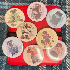 Può includere: Nove bottoni rotondi colorati con vari personaggi dei cartoni animati. I bottoni sono disposti in cerchio su una superficie rossa. I personaggi includono una persona su una sedia a rotelle, una persona con una scatola sulla testa, una persona con un cappello e un bastone, una persona con un berretto e una sciarpa, e una persona con una camicia a quadri.