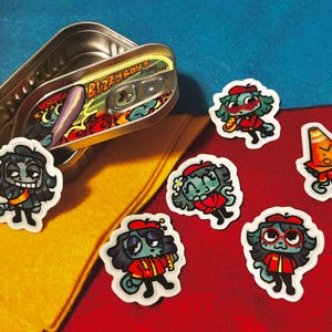 Puede incluir: Una lata abierta de "Bizzy Boys Fish Tin" con una ilustración de dibujos animados. Varias pegatinas de dibujos animados de personajes con chaquetas y boinas rojas están esparcidas alrededor de la lata. El fondo es una mezcla de amarillo, rojo y azul.