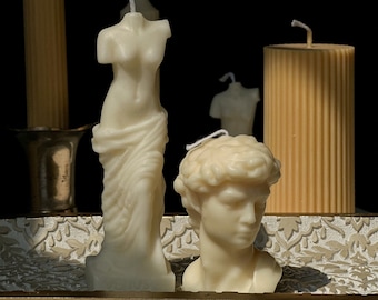 Peut inclure: Trois bougies en cire blanche en forme de sculptures du corps humain. Les bougies sont sur un plateau décoratif à motif floral.