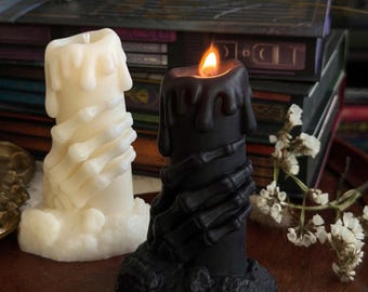 May include: Two decorative candles. One is ivory-colored, the other is black. Both candles are cylindrical with a melted wax appearance and a skeletal hand design wrapped around the base. The black candle is lit with a visible flame.
