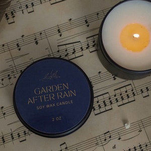 May include: A blue soy wax candle with gold lettering that reads "GARDEN AFTER RAIN" and "SOY WAX CANDLE". The candle is 2 oz and is sitting on a sheet of music.