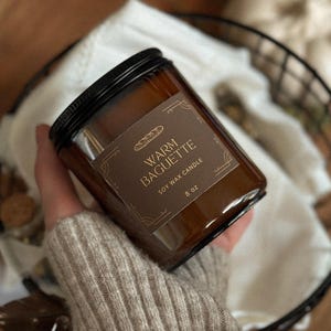 May include: A brown glass jar candle with a black lid. The label reads "Warm Baguette Soy Wax Candle 8 oz." The candle is held in a hand wearing a beige sweater. The background includes a white cloth and a black basket.