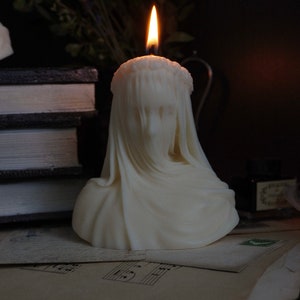 May include: A white candle shaped like a bust of a person wearing a veil and a crown. The candle is lit and the flame is visible. The candle is sitting on a stack of papers with musical notes.