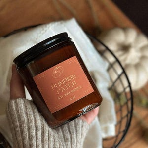 May include: A brown glass jar candle with a black lid, labelled "Pumpkin Patch Soy Wax Candle 8 oz." The label features a pumpkin graphic. The candle is held in a hand wearing a grey sweater, with a basket and a white pumpkin in the background.