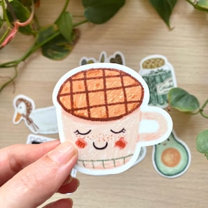 May include: A sticker featuring a cartoon coffee cup with a waffle top. The cup is light pink with a smiling face and rosy cheeks. Other stickers, including an avocado and a jar, are in the background.