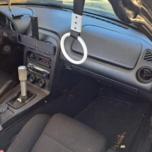 May include: A white plastic ring attached to a black strap hangs from the roof of a black car. The ring is a pull-up handle for the driver.
