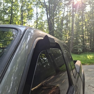 May include: A matte black window visor deflector installed on the upper frame of a gray vehicle door. The accessory follows the contour of the window frame, extending along the top edge to provide a sleek, functional addition to the exterior.