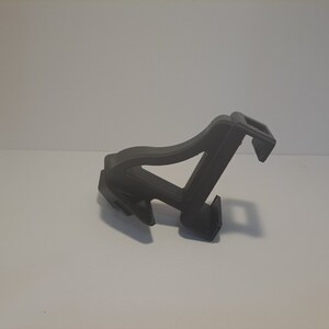May include: A black plastic phone holder with a spring-loaded clamp. The holder is designed to be attached to a surface and can be adjusted to hold a phone at different angles.
