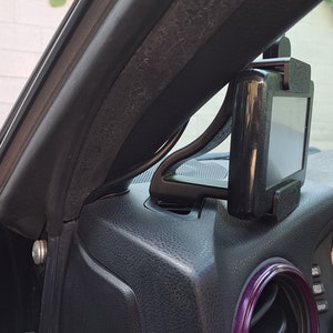 May include: A black car phone mount attached to the dashboard of a car. The mount is designed to hold a phone in a vertical position.