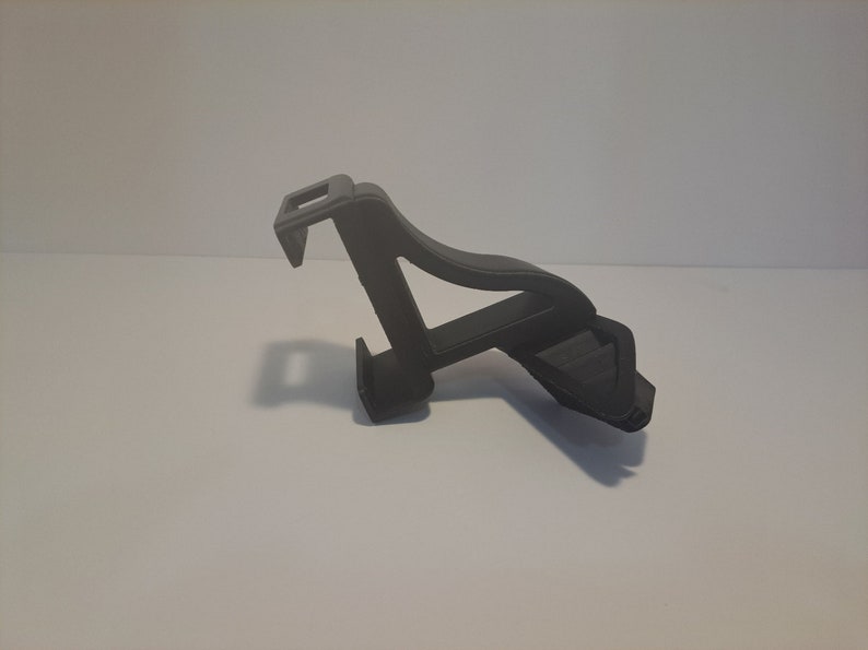 May include: A black plastic phone holder with a clip on the back. The holder is designed to hold a phone in a vertical position.