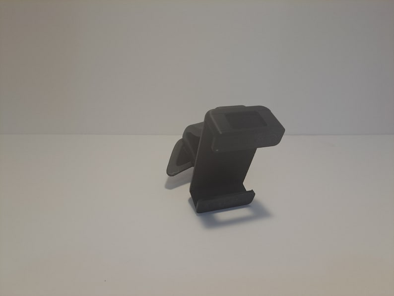 May include: A black plastic phone holder with a clip on the back. The holder is designed to be attached to a car vent.