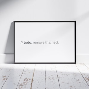 Peut inclure: Impression encadrée avec un cadre noir, affichant le texte "// todo: remove this hack" dans un style minimaliste. L'impression est posée contre un mur blanc et repose sur un sol en bois peint en blanc.