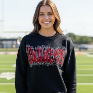 May include: Black sweatshirt with the word "Bulldogs" in red and white rhinestones. The sweatshirt is worn with blue jeans. The background is a green football field.