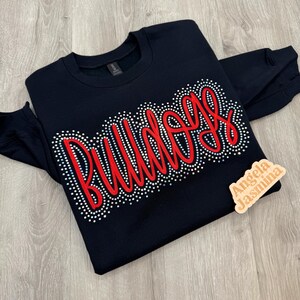 May include: A black sweatshirt with the word "Bulldogs" in red script, outlined with small, sparkling rhinestones. The sweatshirt has long sleeves and a crew neck. A small wooden tag with the text "Angela Jasmina" is on the right side.