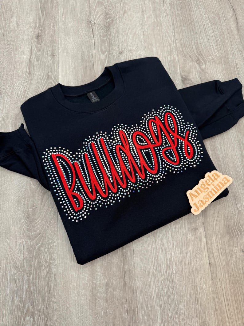 May include: A black sweatshirt with the word "Bulldogs" in red script, outlined with small, sparkling rhinestones. The sweatshirt has long sleeves and a crew neck. A small wooden tag with the text "Angela Jasmina" is on the right side.