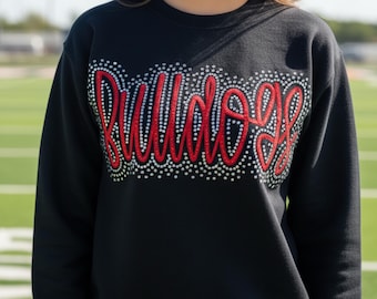 May include: Black sweatshirt with the word "BULLDOGS" in red script, outlined with small white rhinestones. The sweatshirt is a crew neck style. The background is a green field.