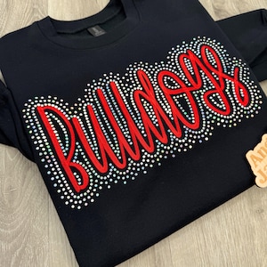 May include: Black crewneck sweatshirt with the word "Bulldogs" in red script, outlined with sparkling rhinestones. The sweatshirt is a solid black color, and the text is the focal point of the design. A wooden tag is in the corner.