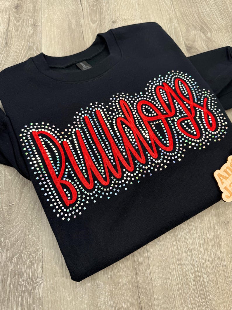 May include: Black crewneck sweatshirt with the word "Bulldogs" in red script, outlined with sparkling rhinestones. The sweatshirt is a solid black color, and the text is the focal point of the design. A wooden tag is in the corner.