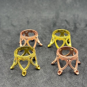 May include: Four small, circular metal display stands arranged in a square formation. Two stands are gold-toned and two are rose gold-toned. Each stand features a crown-like design with heart-shaped cutouts and three small, rounded feet at the base.