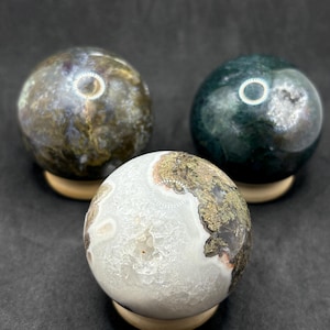 Peut inclure: Trois sphères en pierre polie reposant sur de petits supports en bois. La sphère avant est blanche avec un centre cristallin texturé et des inclusions brunes. La sphère arrière gauche est brun et or, tandis que celle de droite est vert foncé.