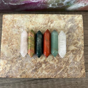 May include: Six polished crystal wands in a row, each with a different colour and pattern. The crystals are arranged on a rectangular, marbled stone surface. Colours include pink, green, black, red, and clear.