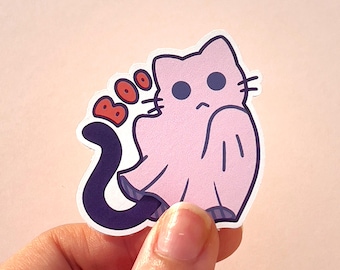 Op de afbeelding: Een roze kat-geest sticker met het woord "BOO" in het rood. De sticker heeft een donkerpaarse staart en wordt tegen een lichtroze achtergrond gehouden. De kat-geest heeft een verdrietige uitdrukking.