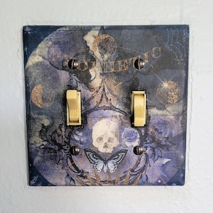 Könnte beinhalten: Ein dekorativer Lichtschalter mit einem dunkelblauen, violetten und goldenen Design. Das Design zeigt einen Schädel, einen Schmetterling und das Wort "Prophethic".