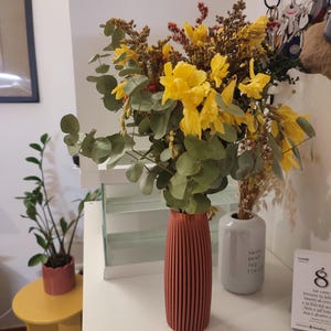 Puede incluir: Un ramo de flores secas amarillas y hojas de eucalipto verdes dispuesto en un jarrón cilíndrico alto y acanalado de color terracota. El arreglo presenta flores amarillas brillantes mezcladas con follaje marrón texturizado y tallos de eucalipto.