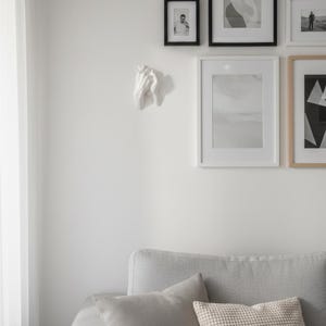 Op de afbeelding: Een witte, gebeeldhouwde hand aan een witte muur. De hand is het middelpunt, met een gedetailleerde textuur. De muur toont ook ingelijste kunstwerken in zwarte en natuurlijke houten lijsten, en een grijze bank met kussens.