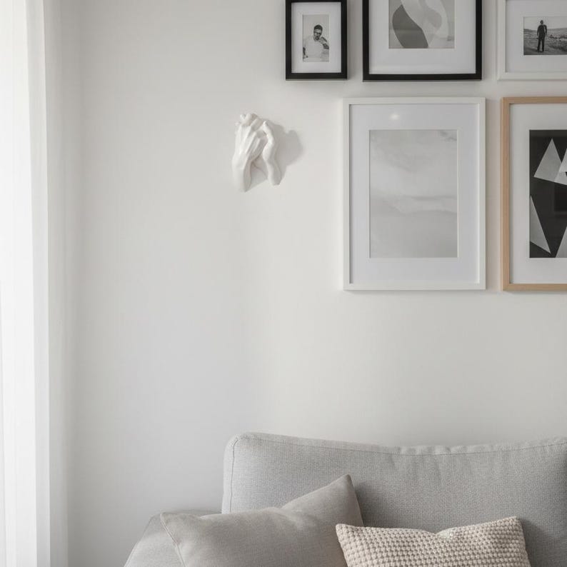 Op de afbeelding: Een witte, gebeeldhouwde hand aan een witte muur. De hand is het middelpunt, met een gedetailleerde textuur. De muur toont ook ingelijste kunstwerken in zwarte en natuurlijke houten lijsten, en een grijze bank met kussens.
