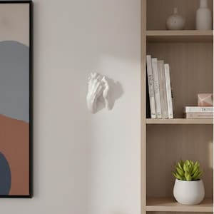 Op de afbeelding: Een witte keramische wandsculptuur die twee in elkaar grijpende handen afbeeldt. De sculptuur is bevestigd aan een effen witte muur. De handen zijn gedetailleerd en lijken in een gebeds- of vasthoudgebaar te zijn. Een decoratief woonaccessoire.