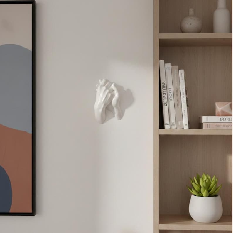 Op de afbeelding: Een witte keramische wandsculptuur die twee in elkaar grijpende handen afbeeldt. De sculptuur is bevestigd aan een effen witte muur. De handen zijn gedetailleerd en lijken in een gebeds- of vasthoudgebaar te zijn. Een decoratief woonaccessoire.