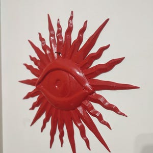 May include: A bright red wall-mounted object shaped like a stylized sun with a central eye. The design features a prominent, almond-shaped eye at the center, surrounded by numerous radiating, wavy, and pointed rays that extend outward in all directions.