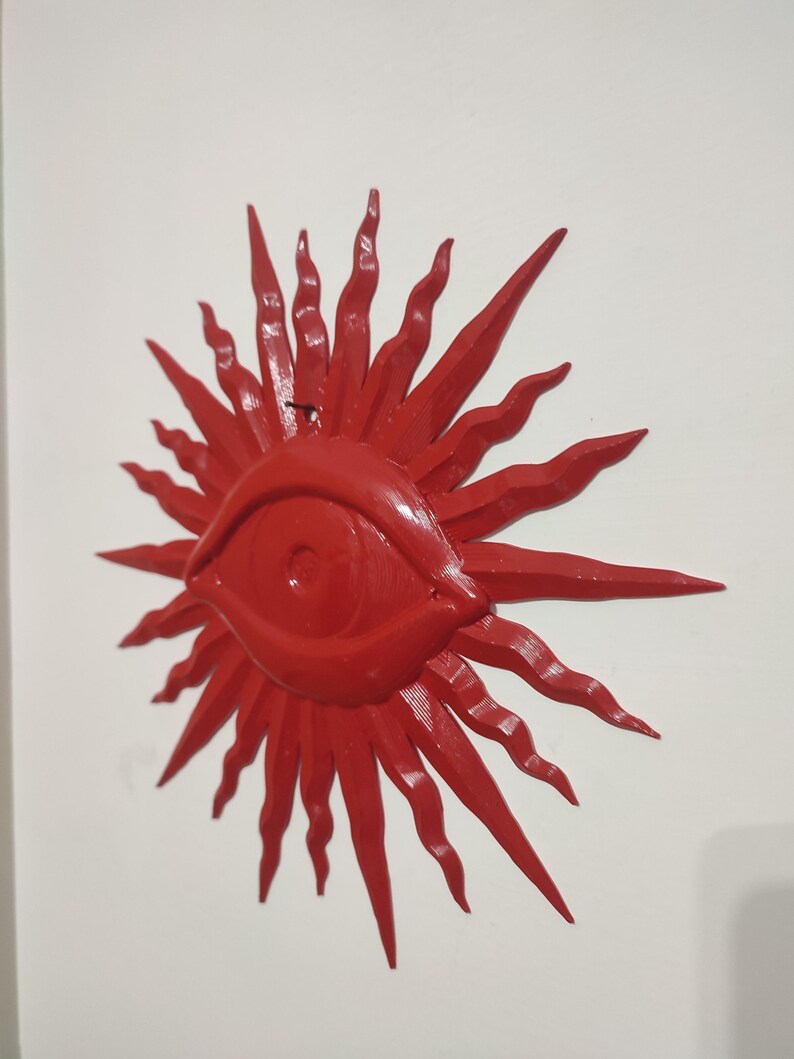 May include: A bright red wall-mounted object shaped like a stylized sun with a central eye. The design features a prominent, almond-shaped eye at the center, surrounded by numerous radiating, wavy, and pointed rays that extend outward in all directions.