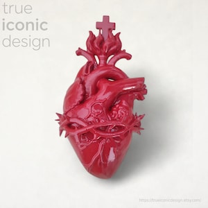 Könnte beinhalten: Glänzend rote Herzskulptur mit Dornenkrone und Kreuz. Der Text "true iconic design" befindet sich oben links. Die Skulptur ist ein religiöses Symbol.