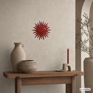 May include: A red wall-mounted sunburst ornament featuring a central eye design. The piece is displayed above a wooden table holding two textured beige vases, a small stone candle holder, and a single red taper candle in a neutral room.