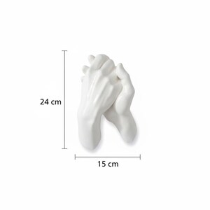 Op de afbeelding: Een witte keramische sculptuur van twee handen die elkaar vasthouden. De sculptuur is 24 cm hoog en 15 cm breed. De handen zijn glad en gedetailleerd, met een glanzende afwerking. Het is een decoratief item.