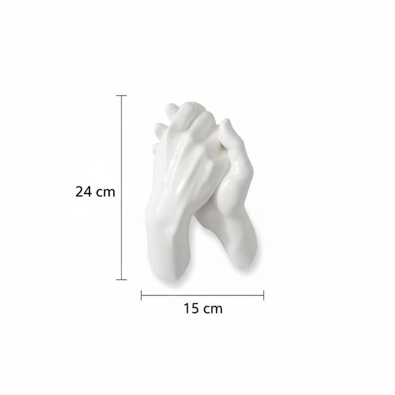 Op de afbeelding: Een witte keramische sculptuur van twee handen die elkaar vasthouden. De sculptuur is 24 cm hoog en 15 cm breed. De handen zijn glad en gedetailleerd, met een glanzende afwerking. Het is een decoratief item.